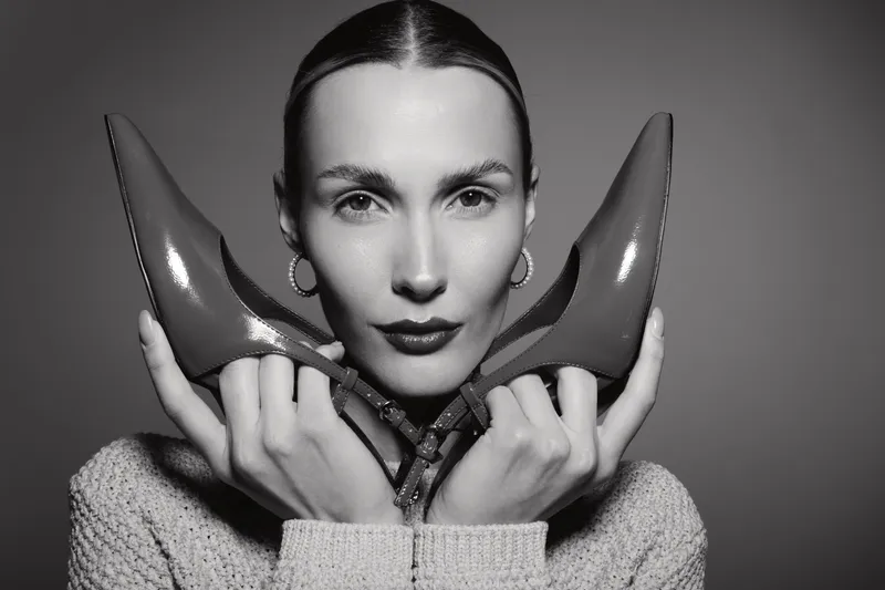 White woman with light skin holding red high heels near face, wearing knitted sweater and earrings. Studio portrait in black and white with fashion concept.