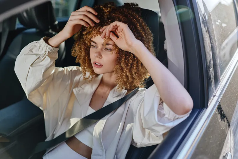 young woman sitting in car wearing seatbelt, curly hair, casual beige jacket, thoughtful expression, warm sunlight, summer driving