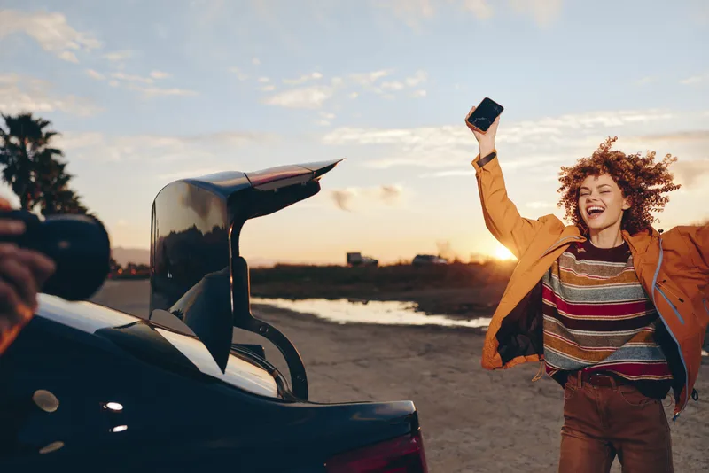 Woman lifestyle at sunset, smiling and holding phone high for a selfie beside a car on an open road, curly hair, orange jacket, warm travel joy outdoors