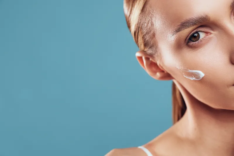 Close-up of woman applying white face cream on cheek with clear skin and natural makeup against blue background, concept of skincare, beauty, and healthy lifestyle.
