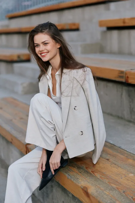 Young businesswoman exuding confidence and style while sitting on wooden stairs in a modern urban setting, dressed in a light-colored suit and smiling radiantly.
