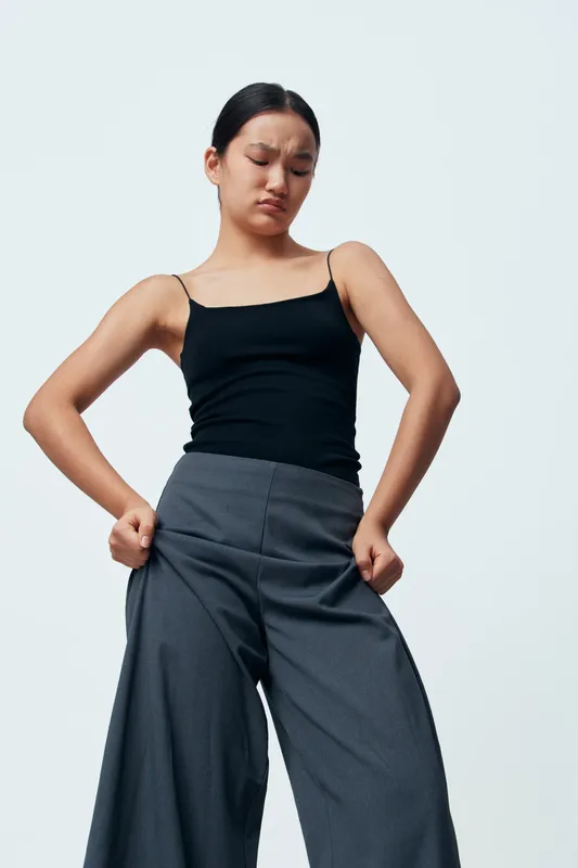 Woman wearing gray pants and black sleeveless top struggling to button pants with frustrated facial expression against plain background. Casual fashion, body image and clothing fit issue.