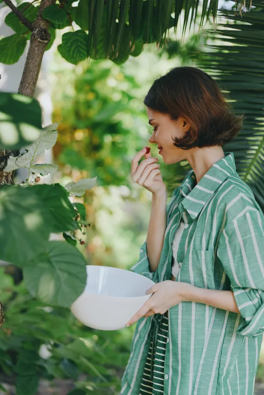A woman in a green striped shirt holds a white bowl as she examines plants in a sunlit garden, capturing a candid moment of outdoor care.