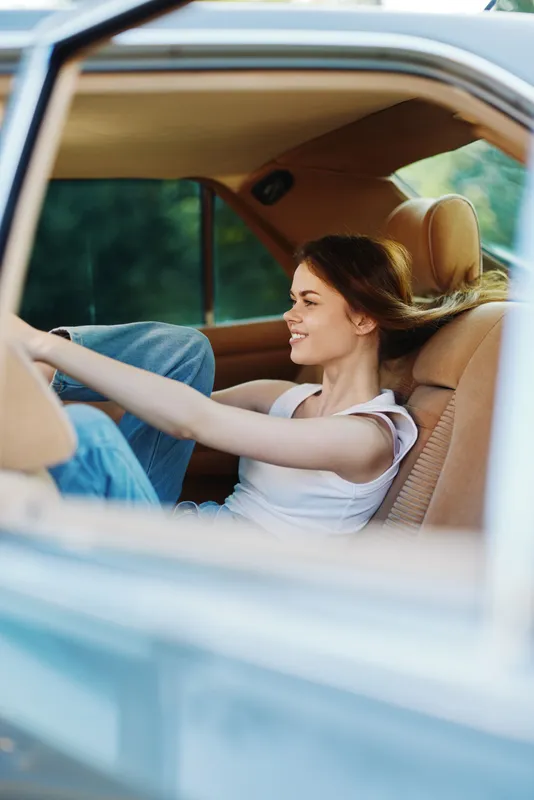 Young woman enjoying a carefree moment in a vintage car, wearing casual attire, smiling through the window with a natural background.