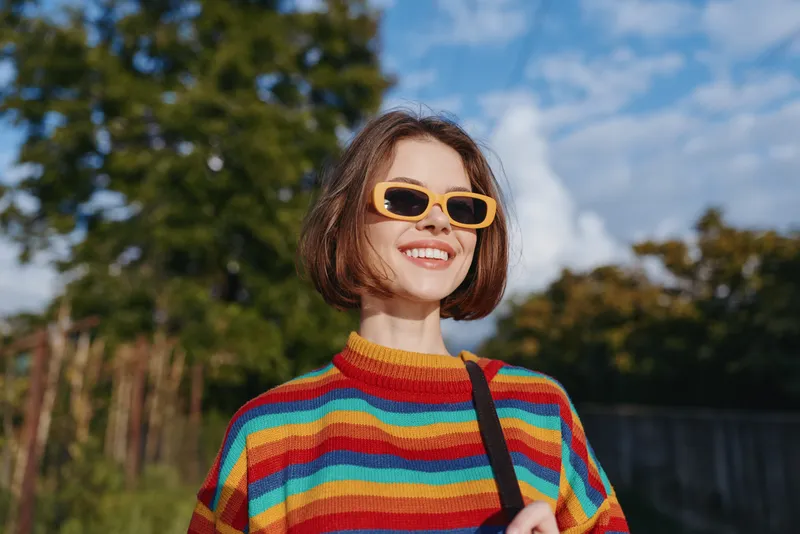 Woman smiling in a rainbow sweater and yellow sunglasses outdoors under blue sky, short hair and casual outfit, carrying a shoulder bag for lifestyle travel and sunny day joy.