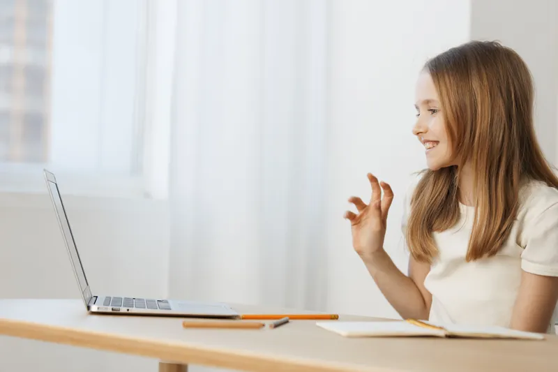 Young girl smiling while video chatting on her laptop, wearing a casual white shirt, in a bright and minimalistic room, evoking joy and connection