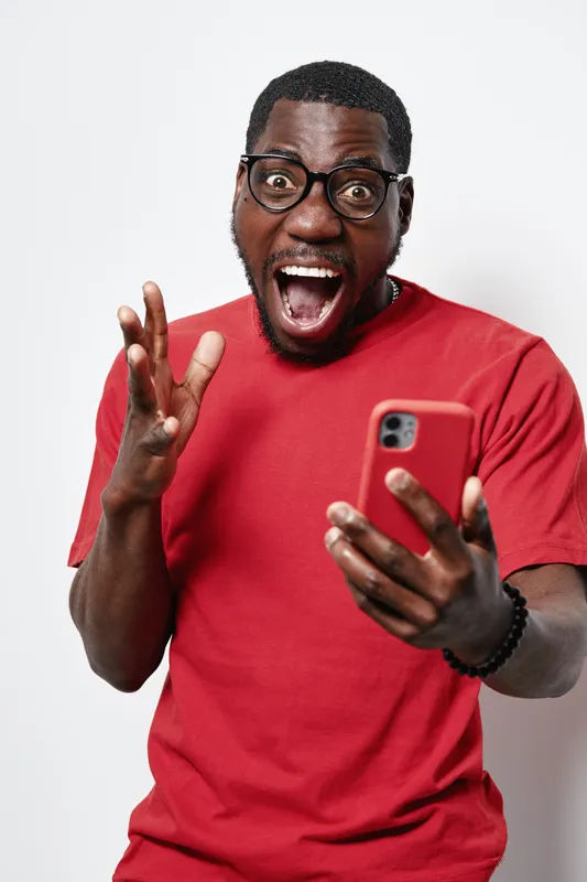 African American man with dark skin and glasses wearing red shirt holding smartphone with surprised expression and open mouth on white background. Casual technology and emotion.