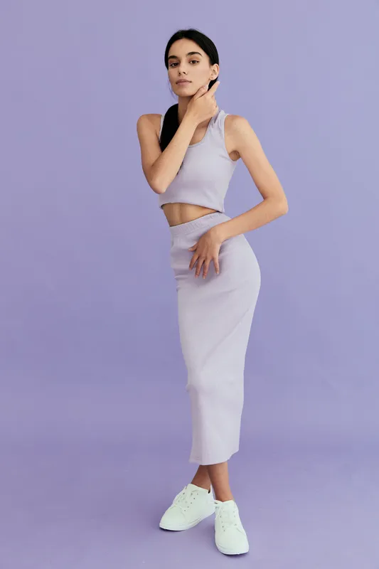 young woman in stylish lavender outfit posing confidently against a purple background with white sneakers, showcasing modern fashion and minimalistic style.