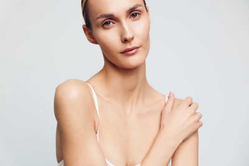 Portrait of young woman with natural skin looking at camera touching shoulder with hand isolated on white background minimal makeup serene expression calm mood.