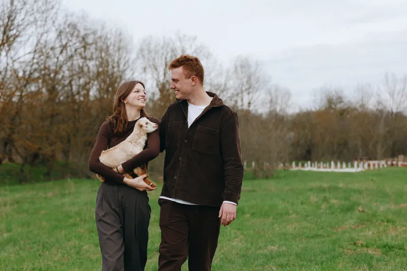 Cheerful couple outdoors, walking in a grassy field with a playful goat kid, warm smiles, relaxed pace, soft breeze, natural colors, sunlight, candid moment, countryside backdrop