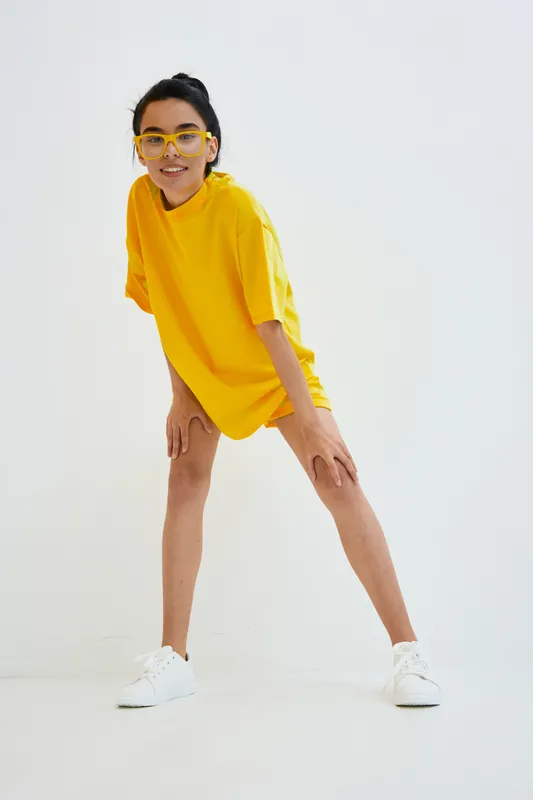 Young woman in bright yellow t-shirt and glasses stretching legs in casual white sneakers against plain white background, showing energy and readiness.