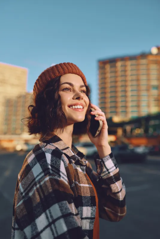 Woman on phone with a warm smile in urban street, beanie and plaid coat at sunset, candid lifestyle shot with golden hour glow, authenticity, mindful living and emotional storytelling.