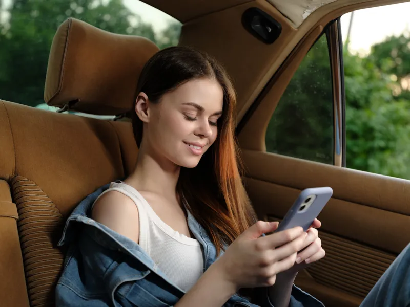 Young woman enjoying her mobile phone while sitting in a vintage car, showcasing casual style and a cheerful smile, with greenery in the background.