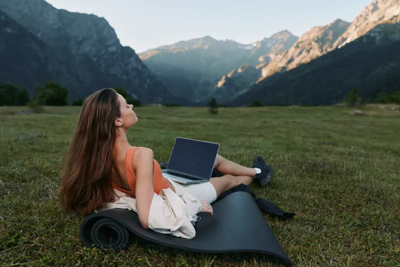 A relaxed woman sits on a yoga mat in a scenic meadow with a laptop resting on her lap, enjoying fresh air and nature while focused on work.