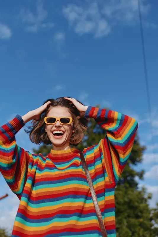 Woman smiling in a rainbow sweater and yellow sunglasses, young adult mid 20s laughing with hands in hair outdoors under blue sky in urban park, joyful casual outfit lifestyle portrait.