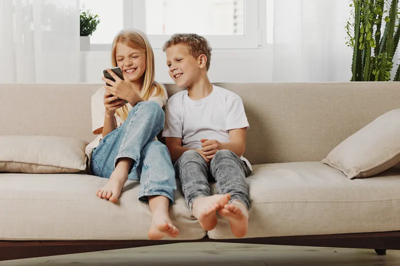 Happy children sitting on sofa indoors, smiling and looking at smartphone together. Casual home environment with natural light, concept of family, technology, and leisure time.
