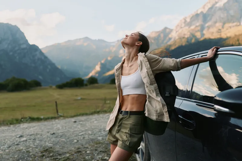 Woman in casual outdoor wear leans against a car amid mountains, enjoying travel and nature, a lifestyle moment of freedom and adventure in luminous scenery.