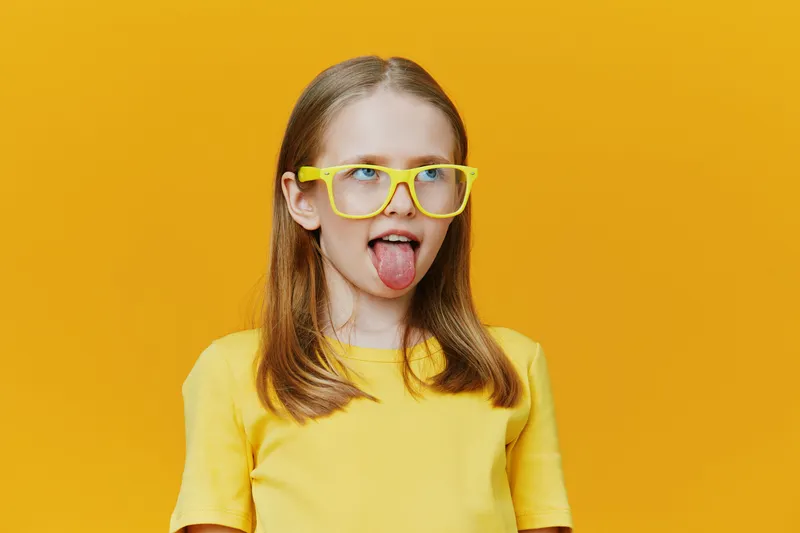 Playful girl making a funny face with yellow glasses, wearing a bright yellow shirt against a vibrant yellow background, exuding joy and energy