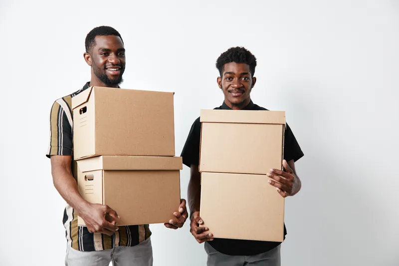 Two young black men with dark skin holding cardboard boxes for moving or delivery. Smiling African American friends standing against white background with casual clothes.