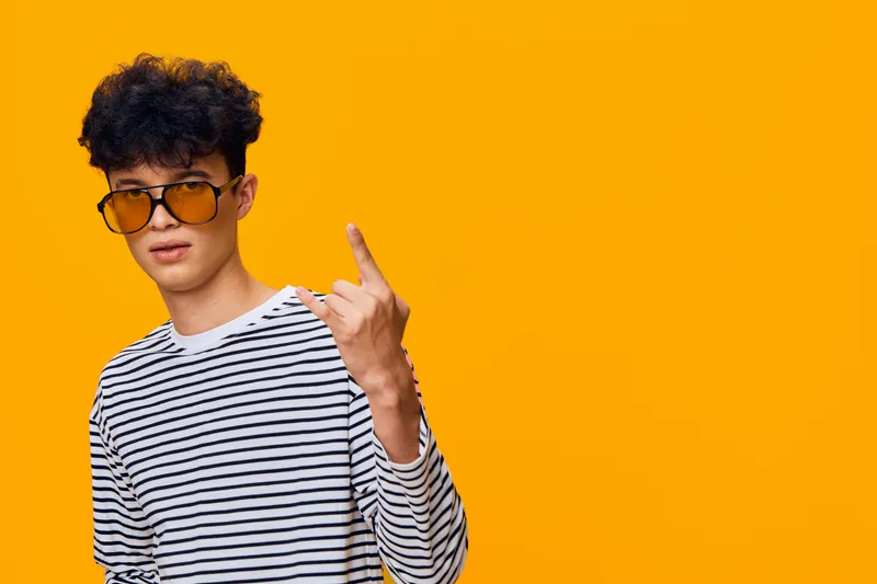 Young man wearing striped shirt and yellow sunglasses making rock sign on bright orange background, showing casual style and confident attitude.
