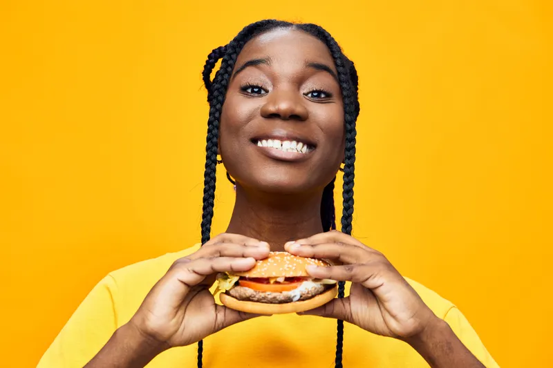 Smiling young woman with braids holding a delicious burger against a bright yellow background, representing joy and healthy eating concepts