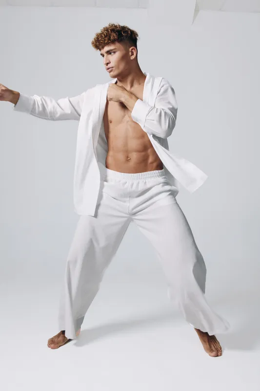 Young man with muscular build practicing martial arts stance wearing loose white pants and open shirt against plain backdrop. Fitness, strength and balance concept with athletic barefoot male.