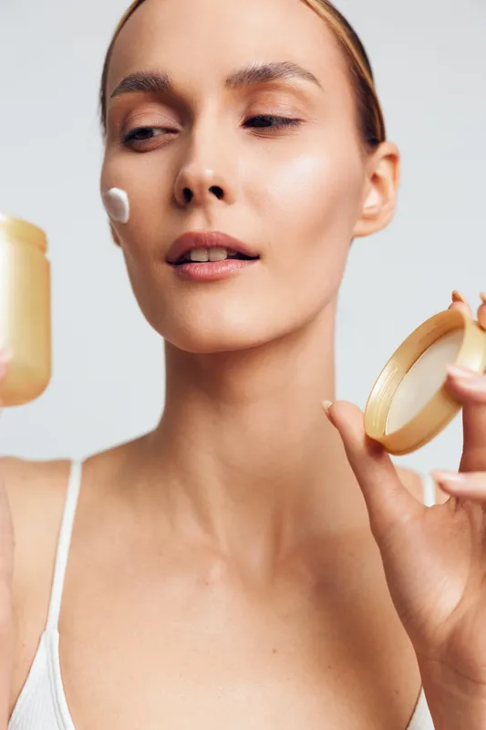 A young woman applies face cream on her cheek holding an open jar in one hand and the lid in the other against a plain background. Concept for skincare and beauty routine.