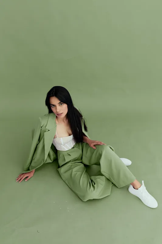 young woman in green suit sitting relaxed on matching green background wearing white sneakers, long dark hair and casual style, simple modern look