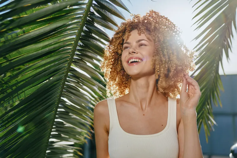 smiling woman with curly hair enjoying sunny tropical outdoors, wearing white tank top framed by green palm leaves and bright sunshine