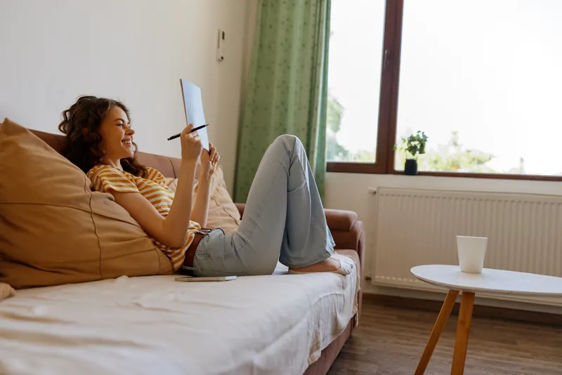Smiling woman with curly hair relaxing on a couch, writing in a notebook, at home. Cozy living space, soft daylight, calm mood, casual creativity and personal downtime.