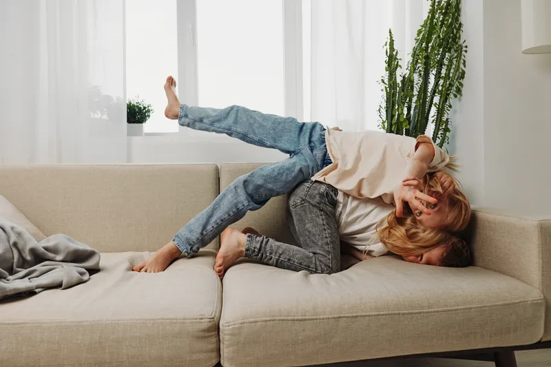 Two playful children in casual clothes wrestle and laugh on a beige sofa in a bright living room with plants in the background. Concept of family fun, childhood joy, and sibling bonding.