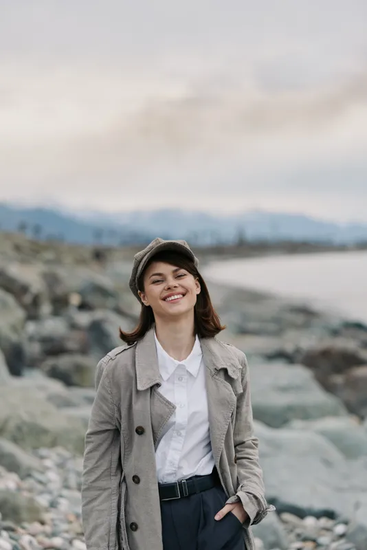 Young woman in patterned coat and hat smiles happily outdoors near rocky shore with blurred water and mountains in background. Concept of nature, fashion, and casual lifestyle.