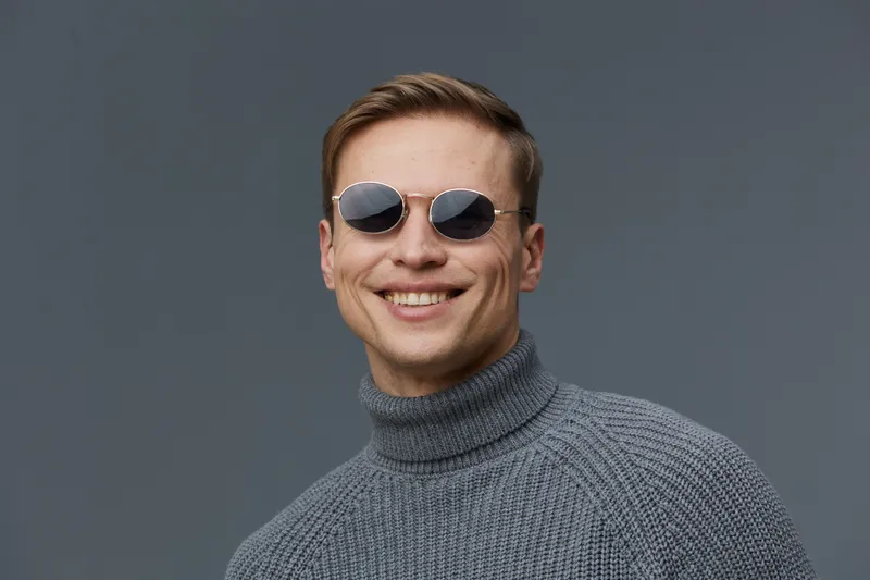 Smiling man wearing stylish sunglasses and a gray turtleneck sweater look cheerful while posing against a plain gray background in a modern studio portrait. People lifestyle concept emphasizing fashio