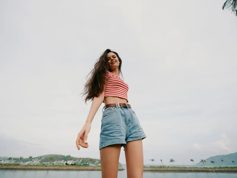 Young woman posing confidently by a breezy seaside setting, wearing a striped crop top and denim shorts with a belt, projecting carefree summer energy and confidence.