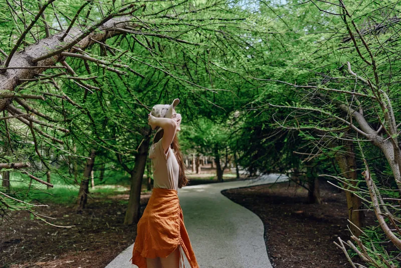 Woman walking on a curved path under dense trees in a green nature park, wearing an orange skirt and sun hat, enjoying summer leisure, motion and a serene outdoor escape.
