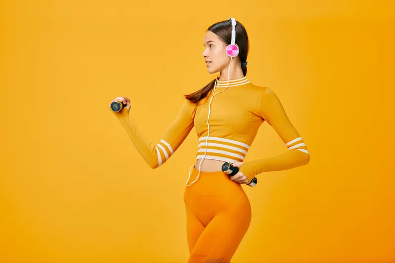 Athletic woman in orange fitness outfit with headphones and dumbbells, posing in a bold orange studio for energetic training and healthy lifestyle visuals.