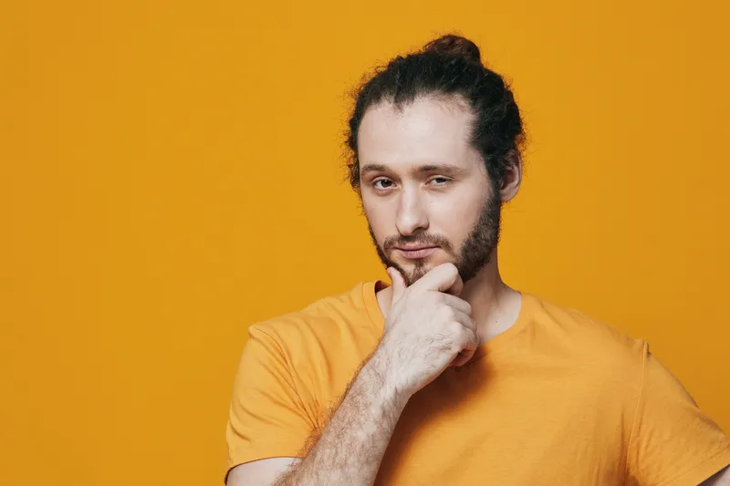 Thoughtful young man with a beard in an orange t shirt against a bright yellow background, contemplating and reflecting with a confident expression