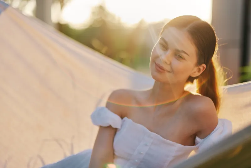 Young woman relaxing in a hammock during golden hour, wearing a light dress, exuding tranquility and happiness while surrounded by nature
