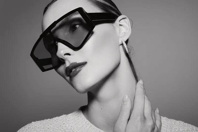 Close-up of a young caucasian woman with pale skin wearing oversized sunglasses and a knitted sweater in a studio setting.