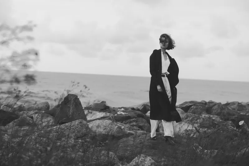 Monochrome scene of a woman standing on rugged rocks by the sea, wearing a long coat and flowing hair, capturing a windy coastal landscape with open horizon and dramatic mood.