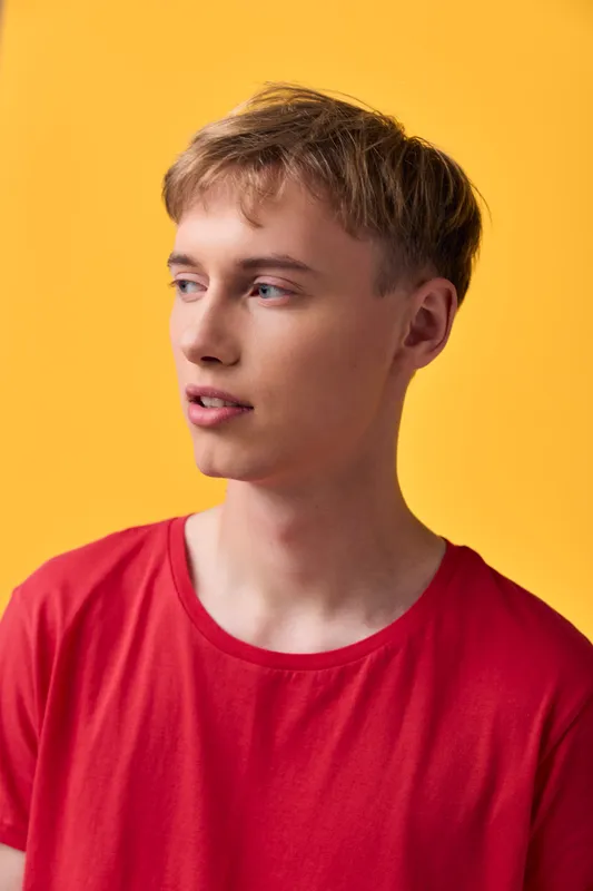 Young man in a red shirt stands against a bold yellow backdrop in a studio, showcasing casual fashion, modern style, close up detail, and a friendly expression with clean skin for lifestyle use