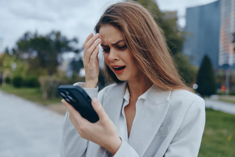 Business woman expressing frustration while using a smartphone outdoors in a city setting. She wears a light blazer and has long hair, showcasing a professional appearance.