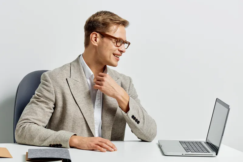 Smiling man in smart casual attire working on laptop at modern office desk with notepad and glasses, expressing confidence and professionalism. Studio shot with neutral background. People working conc