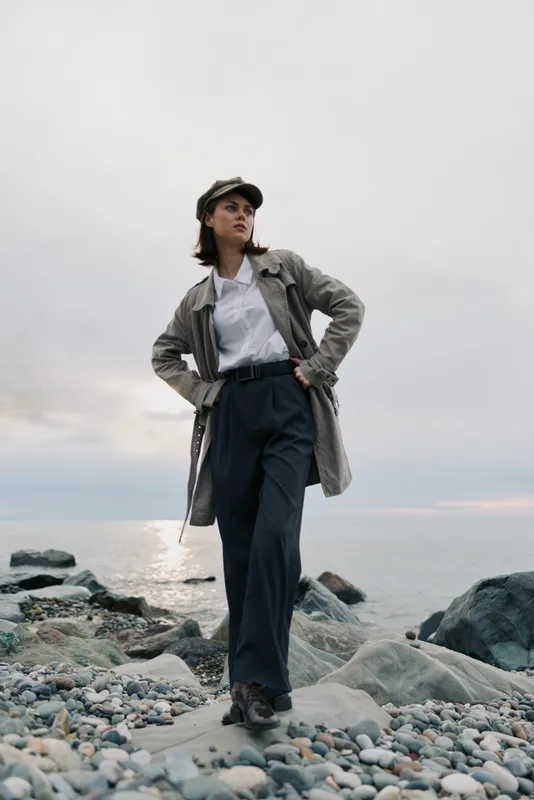 Confident young woman wearing a trench coat and cap stands on a rocky beach with hands on hips. Overcast sky and calm sea create a moody, contemplative atmosphere.