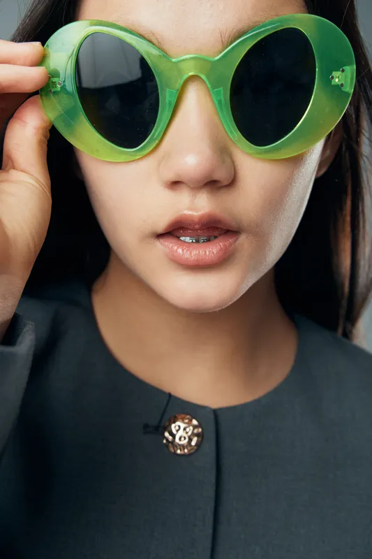Woman wearing large green sunglasses with dark lenses closeup, showing natural lips and black hair with gray clothing and gold button on collar for fashion look.