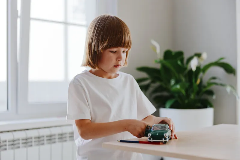 Young boy playing with a toy car at home, showcasing concentration and joy in a bright, minimalist environment with green plants.