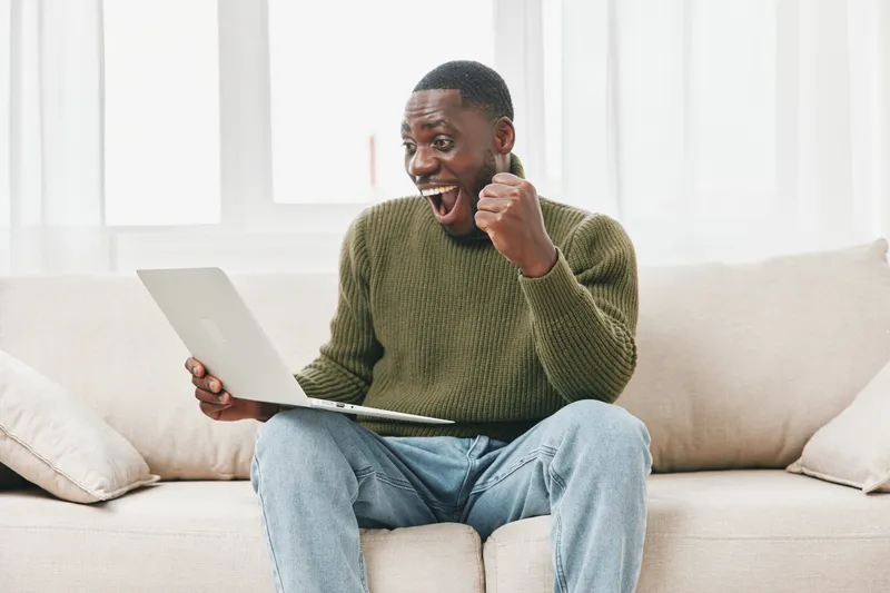 Joyful young African man celebrating success while working on a laptop at home, showcasing excitement in a cozy setting with soft natural light