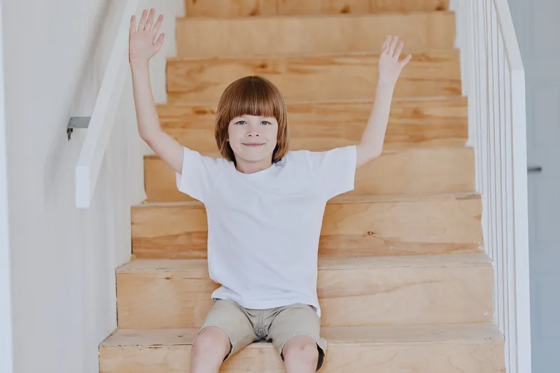 Cheerful boy enjoying playtime on wooden stairs, wearing a casual white t-shirt and shorts, spreading his arms joyfully for a fun and carefree atmosphere.