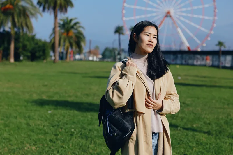 Outdoor scene with a woman in a beige coat standing on a green park, a backpack slung over her shoulder, under a sunny sky with a distant ferris wheel