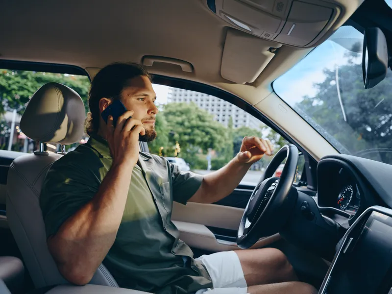 A man driving a car while talking on a mobile phone inside a sunlit vehicle, focused on the road, gripping the wheel, and maintaining a calm, attentive posture.